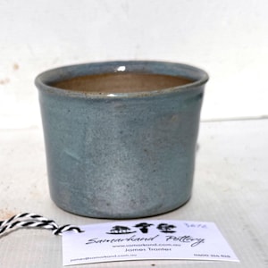 May include: A light blue ceramic planter with a brown rim. The planter is handmade and has a subtle speckled glaze. The planter is labelled "Sun-Hand Pottery" with a website address and the name "James Trotter".