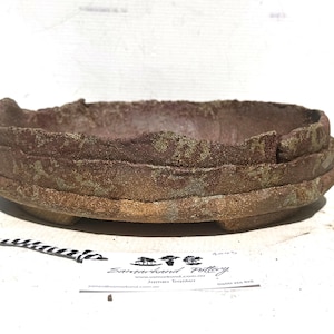 May include: A handcrafted, round, ceramic bonsai pot with a layered design. The pot is primarily reddish-brown with hints of green and a textured surface. The pot has a low profile and is suitable for bonsai trees.