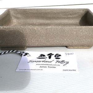 Może przedstawiać: Prostokątny, jasnobrązowy ceramiczny pojemnik na bonsai z czterema małymi nóżkami. Pojemnik ma szorstką, fakturowaną powierzchnię. Do pojemnika przymocowana jest biała etykieta z czarnym tekstem. Etykieta zawiera napis "Samarkand Pottery", "www.samarkand.com.au", "James Tranter", "james@samarkand.com.au" oraz "0400 214 815". Na etykiecie widnieje również numer "3400".