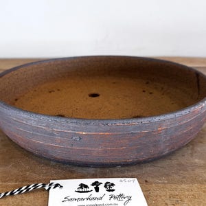 May include: A shallow, round ceramic bonsai pot with a dark, textured exterior featuring horizontal ridges and a warm, reddish-brown interior base. The pot has visible drainage holes and a white tag attached with black and white string.
