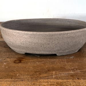 May include: A shallow, oval-shaped ceramic bonsai pot with a speckled, matte grey finish. The bowl features a wide, open top and a smooth, rounded rim, with a slightly darker interior surface and a sturdy base designed for plant cultivation.