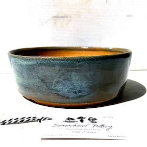 May include: A handcrafted ceramic bowl with a wide, shallow form. The outside has a mottled blue-green glaze, and the inside has a warm, earthy brown hue. The bowl rests on a matching base, ideal for serving or display.