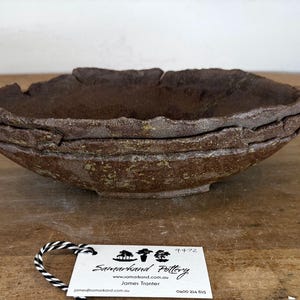 May include: A stack of three dark brown ceramic bowls with a rough, textured surface and uneven, organic edges. A white rectangular tag is attached with black and white string, featuring the text Samarkand Pottery, a website, and contact details.
