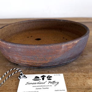 May include: A round, shallow ceramic bonsai pot with a textured, dark brown and reddish-brown exterior finish. The interior is a lighter, speckled tan color with visible drainage holes. A white tag with black text is attached by a black and white string.