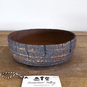 May include: A round ceramic bowl with a dark, heavily textured exterior featuring deep cracks and golden brown highlights. The interior is a smooth, dark brown clay. A white tag with the text Samarkand Pottery is attached by a black and white string.