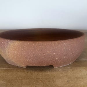 May include: A round, shallow ceramic bonsai pot with a textured, speckled terracotta-colored surface. The exterior features subtle variations in tone, ranging from reddish-brown to muted golden hues, with a small notch at the base.