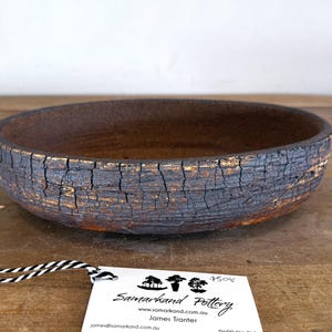 May include: A shallow, round ceramic bowl with a dark, heavily cracked exterior texture revealing golden-brown undertones. The interior is smooth and dark brown. A white tag attached with black and white string reads Samarkand Pottery.