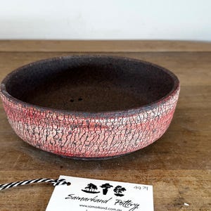 May include: A round ceramic bowl with a dark brown interior and a textured exterior featuring a red and white crackle glaze finish. A white rectangular tag with black text reading Samarkand Pottery is attached by a black and white string.