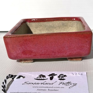 May include: A square, red bonsai pot with a light brown interior. The pot has four small feet and is made of ceramic.