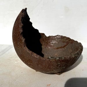 May include: A dark brown, textured ceramic bowl with a jagged, uneven rim. The vessel features a deep, rounded basin and a rough, earthy exterior finish. The bowl is supported by small, subtle feet at the base, creating a distinct, asymmetrical silhouette.