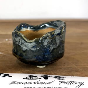 May include: A small, hand-formed ceramic bowl with an irregular, wavy rim. The exterior features a mottled glaze in shades of deep blue, grey, and black, while the interior reveals a smooth, light tan finish. A white tag with black text reads Samarkand Pottery.