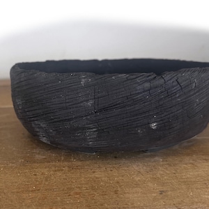 May include: A round, shallow bowl with a matte black finish and a deeply textured, charred wood grain exterior. The surface features prominent, linear cracks and grooves that wrap around the circumference of the vessel, creating a rugged, dark appearance.