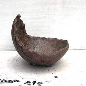 May include: A handcrafted, brown, ceramic bowl with a unique, organic shape. The bowl has a rough, textured surface and sits on three small feet. The rim is uneven, adding to its rustic charm. Ideal for decorative purposes or as a small planter.