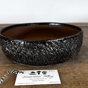 May include: A round, shallow ceramic bowl with a dark, textured, glossy exterior and a smooth, matte brown interior. A white rectangular tag is attached with black and white string, featuring the text Samarkand Pottery, james@samarkand.com.au, James Tranter, 4518, and 0400 214 615.