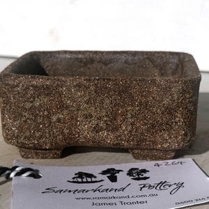 May include: A small, rectangular, brown ceramic planter with a textured surface. The planter has a square shape with a slightly inward-sloping rim and sits on four small feet. A tag with the words "Samarkand Pottery" is attached.