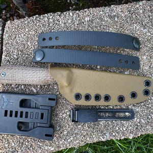 May include: A tactical knife set with a tan leather sheath, black belt loops, and a black mounting system. The knife has a textured handle. The set includes a black belt clip. The knife and accessories are displayed on a stone surface.