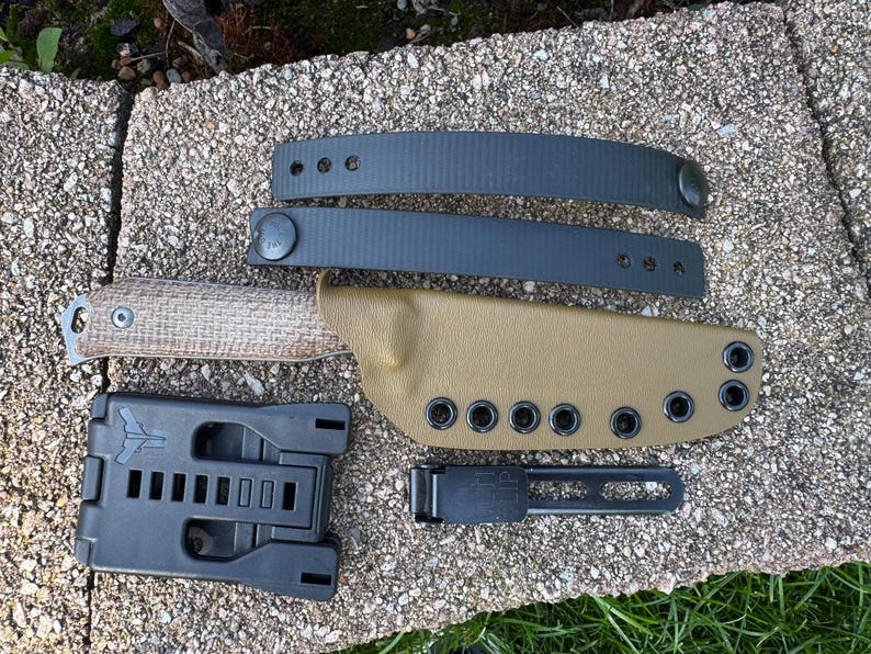 Peut inclure: Un ensemble de couteau tactique avec un &eacute;tui en cuir beige, des passants de ceinture noirs et un syst&egrave;me de montage noir. Le couteau a un manche textur&eacute;. L'ensemble comprend un clip de ceinture noir. L'ensemble est pos&eacute; sur une surface en pierre.