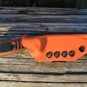 Puede incluir: Un cuchillo negro y naranja con un mango texturizado y una funda de cuero naranja brillante con ojales negros.