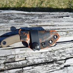 May include: A black and orange knife sheath with a black nylon strap and snap closure. The sheath has multiple black eyelets.