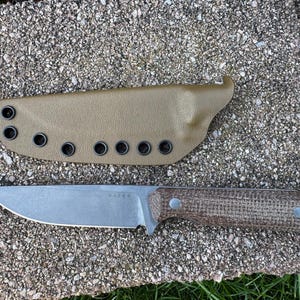 Peut inclure: Un couteau &agrave; lame fixe avec une lame argent&eacute;e et un manche marron, &agrave; c&ocirc;t&eacute; d'un &eacute;tui beige. Le couteau porte le mot "KIZER" grav&eacute; sur la lame. L'&eacute;tui comporte plusieurs &oelig;illets.