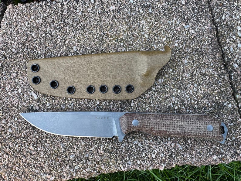 Peut inclure: Un couteau &agrave; lame fixe avec une lame argent&eacute;e et un manche marron, &agrave; c&ocirc;t&eacute; d'un &eacute;tui beige. Le couteau porte le mot "KIZER" grav&eacute; sur la lame. L'&eacute;tui comporte plusieurs &oelig;illets.