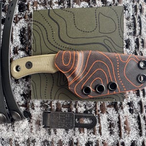 May include: A small, tan-handled knife with a brown and orange patterned sheath, black straps, and a black clip. The sheath has a topographic design. The knife and accessories are on a green card with a topographic design.