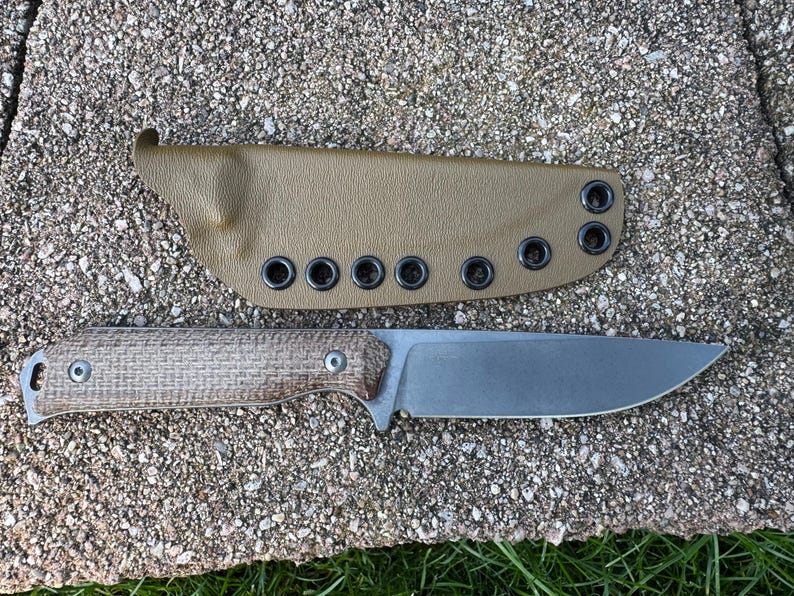 Peut inclure: Un couteau &agrave; lame fixe avec un manche textur&eacute; et une lame argent&eacute;e, accompagn&eacute; d'un &eacute;tui beige. L'&eacute;tui comporte plusieurs &oelig;illets. Le couteau et l'&eacute;tui sont pr&eacute;sent&eacute;s sur une surface textur&eacute;e semblable &agrave; de la pierre.