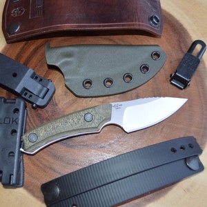 K&ouml;nnte beinhalten: Ein gr&uuml;nes und braunes Messer mit schwarzem Griff und silberner Klinge. Das Messer liegt auf einer Holzoberfl&auml;che mit einer schwarzen Lederscheide, einer schwarzen Plastikscheide und einem schwarzen Plastikclip.