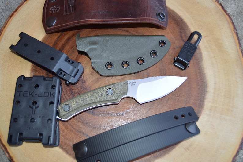 K&ouml;nnte beinhalten: Ein gr&uuml;nes und braunes Messer mit schwarzem Griff und silberner Klinge. Das Messer liegt auf einer Holzoberfl&auml;che mit einer schwarzen Lederscheide, einer schwarzen Plastikscheide und einem schwarzen Plastikclip.