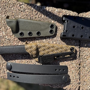 Puede incluir: Un cuchillo negro y marrón con un mango texturizado y una funda negra. El cuchillo está sobre una superficie marrón y gris. Hay una funda verde y un clip de plástico negro al lado del cuchillo.