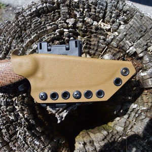 May include: A tan leather knife sheath with multiple black eyelets and a black plastic clip. The sheath is attached to a brown wooden surface.