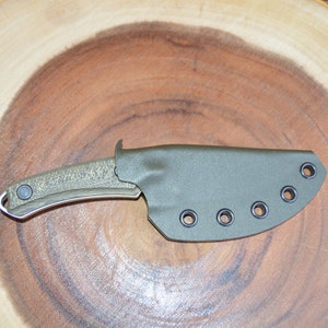 K&ouml;nnte beinhalten: Ein gr&uuml;nes Messer mit einem strukturierten Griff und einer schwarzen Scheide mit sechs schwarzen &Ouml;sen. Das Messer liegt auf einer Holzoberfl&auml;che.