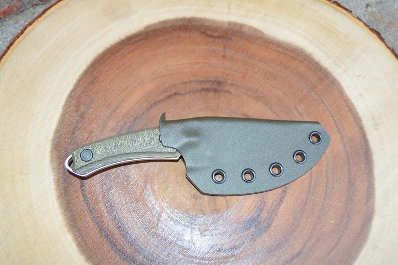 K&ouml;nnte beinhalten: Ein gr&uuml;nes Messer mit einem strukturierten Griff und einer schwarzen Scheide mit sechs schwarzen &Ouml;sen. Das Messer liegt auf einer Holzoberfl&auml;che.