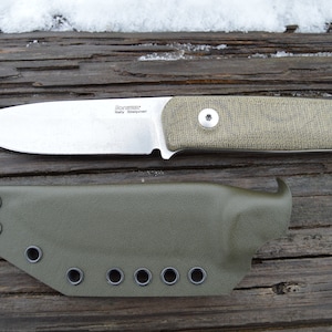 May include: A fixed-blade knife with a silver blade and a green handle, accompanied by a matching green sheath. The knife has the text "LionSteel Italy Sleipner" on the blade. The knife and sheath are displayed on a weathered wooden surface.