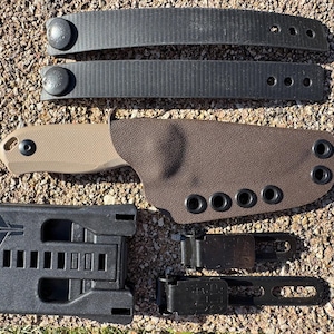 Può includere: Un set di attrezzatura tattica, tra cui un fodero per coltello marrone, un sistema di montaggio nero, due cinghie nere con bottoni a pressione e due clip da cintura nere. Il fodero del coltello è marrone con occhielli neri. Gli oggetti sono disposti su una superficie strutturata marrone chiaro.