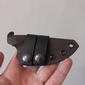 K&ouml;nnte beinhalten: Eine braune Ledertasche f&uuml;r ein Messer mit zwei schwarzen Gummib&auml;ndern und silbernen Druckkn&ouml;pfen. Die Tasche hat vier schwarze &Ouml;sen.