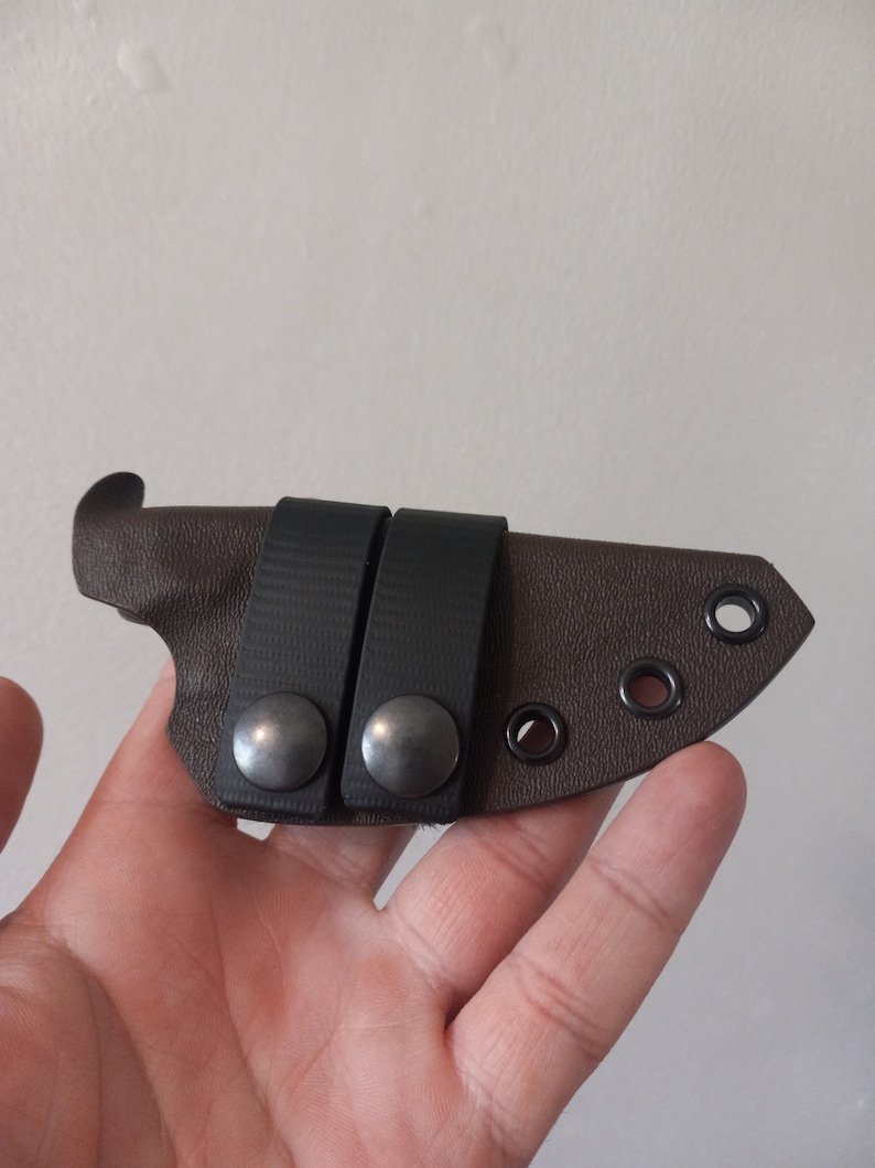 K&ouml;nnte beinhalten: Eine braune Ledertasche f&uuml;r ein Messer mit zwei schwarzen Gummib&auml;ndern und silbernen Druckkn&ouml;pfen. Die Tasche hat vier schwarze &Ouml;sen.