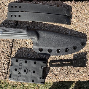 May include: Black knife sheath with six snap closures and a black knife with a textured handle. The sheath is labelled "TEK-LOK" and "BLADE-TECH" with the text "MADE IN USA".