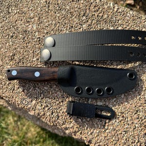 May include: A black leather knife sheath with a brown wooden handle knife inside. The sheath has six metal eyelets and a black plastic clip. A black nylon strap with two metal snaps is also included.