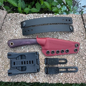 May include: A knife set with a purple handle and a red sheath. The set includes a black belt clip, two black straps, and a black mounting system. The knife handle has a gold-colored eyelet.