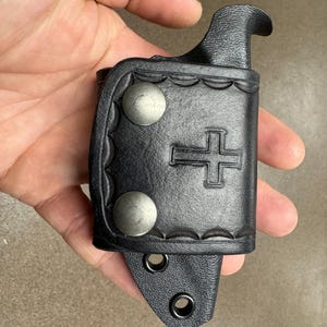 K&ouml;nnte beinhalten: Schwarze Ledertasche f&uuml;r ein Messer mit Kreuzdesign und zwei silbernen Druckkn&ouml;pfen. Die Tasche hat einen G&uuml;rtelschlaufe und einen Druckknopfverschluss.