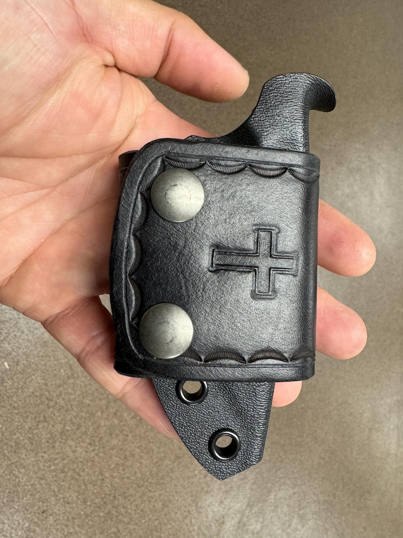 K&ouml;nnte beinhalten: Schwarze Ledertasche f&uuml;r ein Messer mit Kreuzdesign und zwei silbernen Druckkn&ouml;pfen. Die Tasche hat einen G&uuml;rtelschlaufe und einen Druckknopfverschluss.