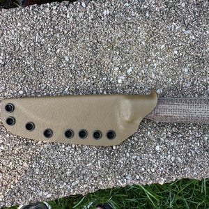 Peut inclure: Un &eacute;tui de couteau en cuir beige avec des &oelig;illets noirs et un couteau &agrave; manche textur&eacute;. Le manche du couteau pr&eacute;sente un motif tiss&eacute;. L'&eacute;tui est con&ccedil;u pour maintenir le couteau en s&eacute;curit&eacute;. Le couteau et l'&eacute;tui reposent sur une surface textur&eacute;e.