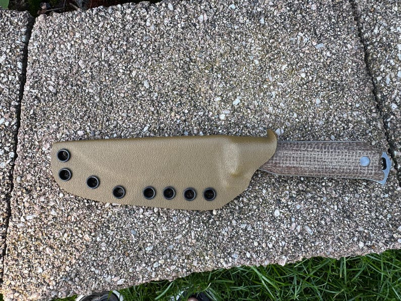 Peut inclure: Un &eacute;tui de couteau en cuir beige avec des &oelig;illets noirs et un couteau &agrave; manche textur&eacute;. Le manche du couteau pr&eacute;sente un motif tiss&eacute;. L'&eacute;tui est con&ccedil;u pour maintenir le couteau en s&eacute;curit&eacute;. Le couteau et l'&eacute;tui reposent sur une surface textur&eacute;e.