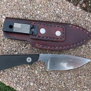 May include: A fixed-blade knife with a black handle and a silver blade, next to a brown leather sheath. The sheath has a black clip with an American flag design. The knife and sheath are on a textured surface.