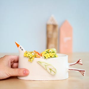 A hand holds the white ceramic bird planter filled with yellow and orange flowers against a light blue background.