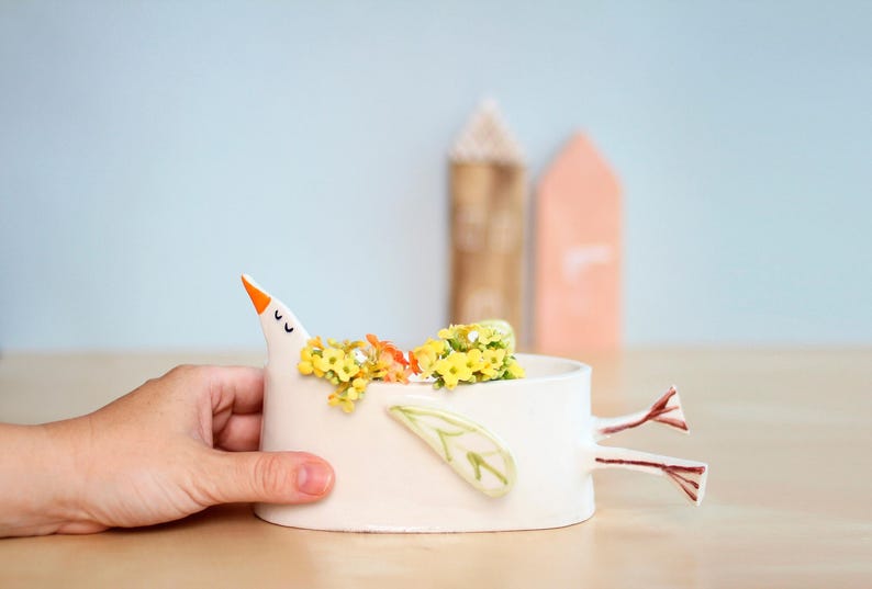 A hand holds the white ceramic bird planter filled with yellow and orange flowers against a light blue background.