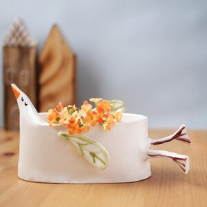 The white ceramic bird planter filled with orange flowers sits on a wooden surface with two wooden house figurines in the background.