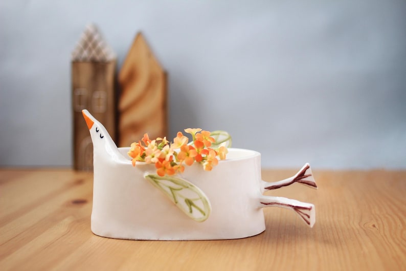 The white ceramic bird planter filled with orange flowers sits on a wooden surface with two wooden house figurines in the background.