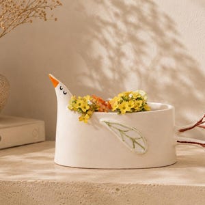 A white ceramic bird planter filled with yellow flowers, resting on a beige surface with a book and vase in the background.